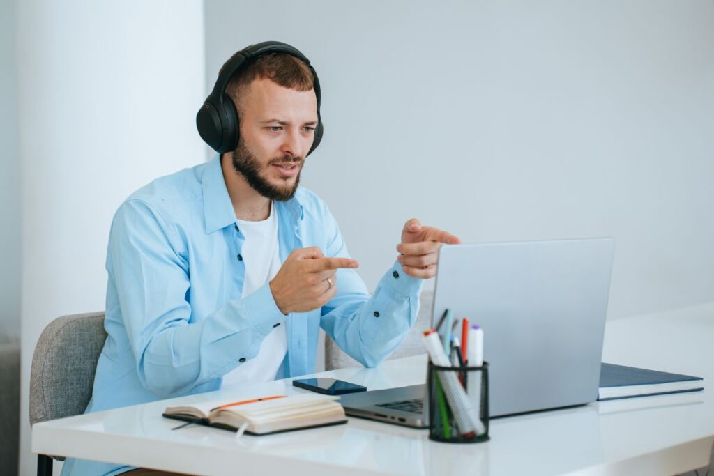 You can do it! Bearded handsome young business man sitting at desk makes video call talks with partner gesturing, using headphones. Purposeful male entrepreneur at video conference giving instructions.