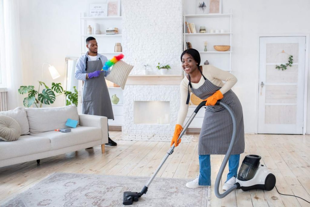 Happy black woman vacuuming floor while her boyfriend dusting cushion at their apartment, full length. Millennial African American couple cleaning home together, copy space