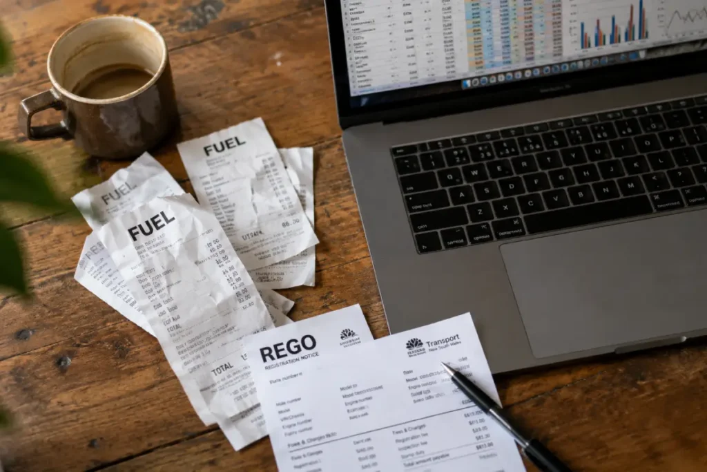 A top-down view of a kitchen table covered in a household budget spreadsheet, fuel receipts, and a vehicle registration notice.